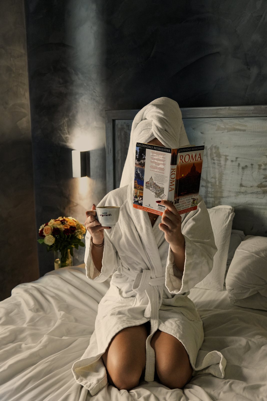 A guest in a white bathrobe and towel sits on the bed with an espresso and a Roma guidebook, lit by the wall sconce.